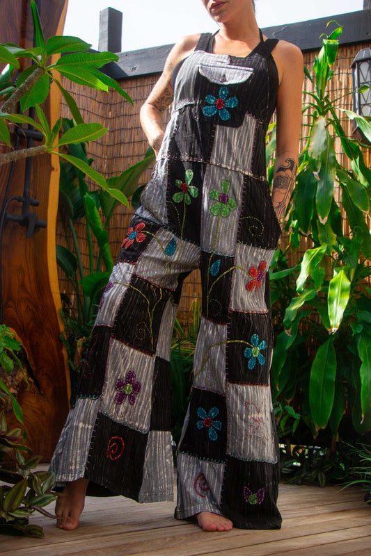A woman wearing embroidered flower patchwork overalls with palazzo legs, standing barefoot on a wooden deck surrounded by lush green plants