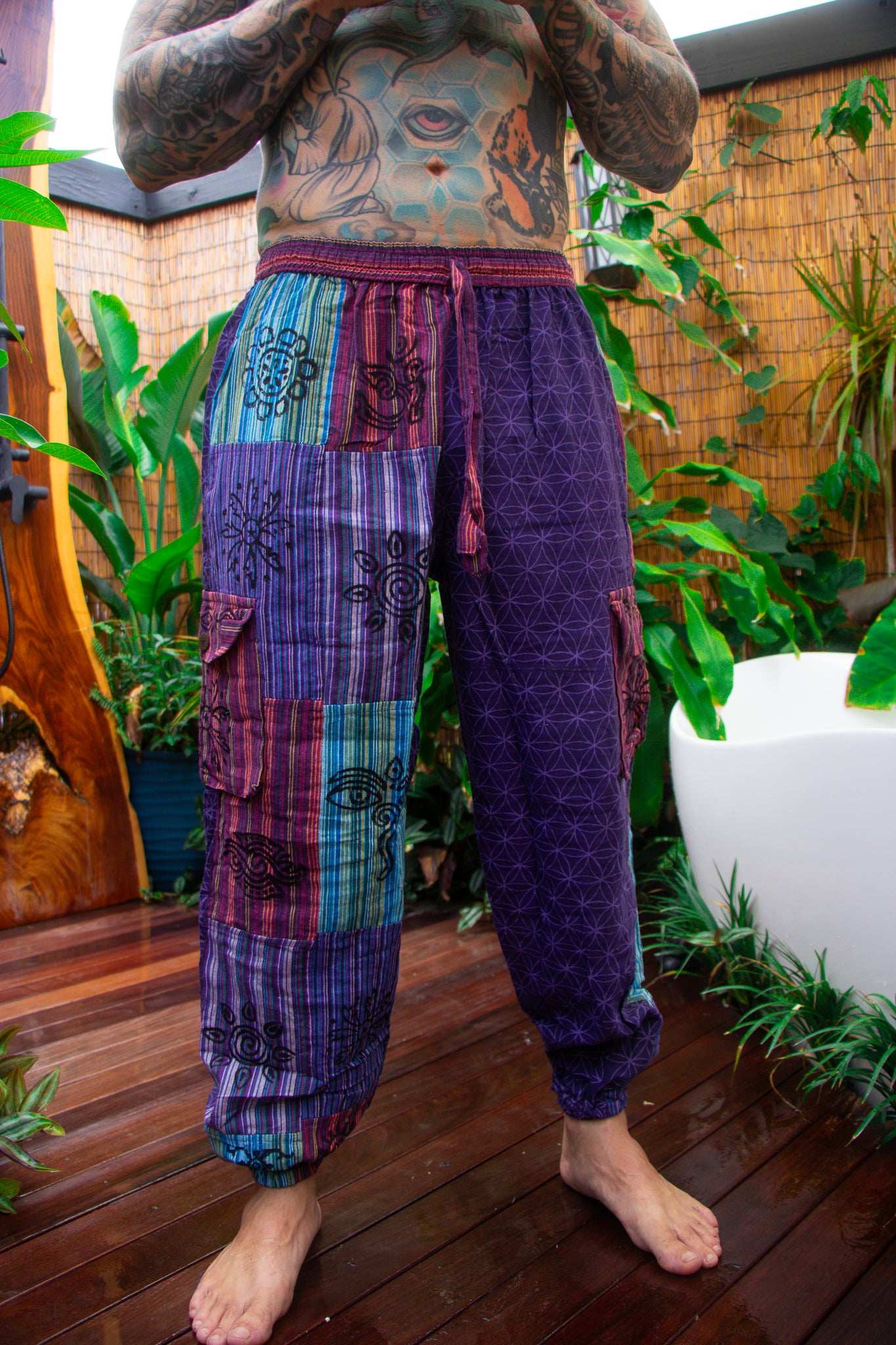 A person wearing loose patchwork harem-style pants with purple, blue, and teal panels, standing barefoot on a wooden deck with plants in the background.